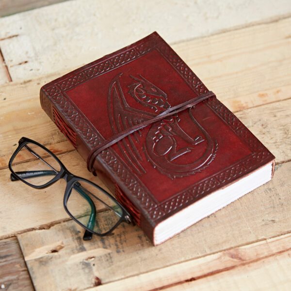 A closed Celtic Sitting Dragon Leather Journal with a strap, embossed in red leather, rests on a wooden surface beside black-framed glasses. The intricate dragon design adds an air of mystique to the scene.