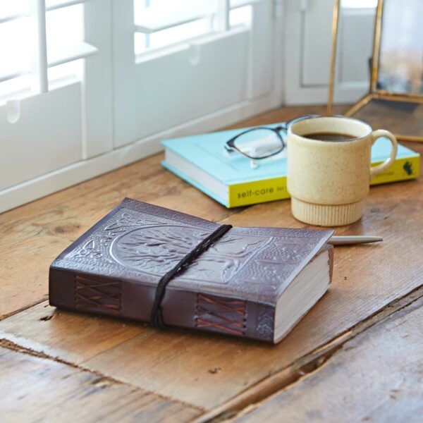 The Tree of Life Leather Journal rests on a wooden table beside a coffee mug, glasses, and a pen. Nearby lies "self-care," its light blue cover adding calm under the window's shuttered light.