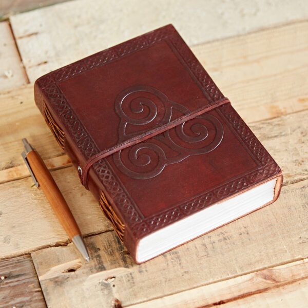 The Celtic Triskelion Symbol Leather Journal with a brown leather cover lies closed next to a wooden pen on a wooden surface.
