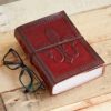 A Fleur de Lis Leather Journal with a red design and wraparound strap lies on a wooden surface beside eyeglasses.