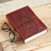 A closed Ganesh Leather Journal with an embossed design sits elegantly beside a pair of black-framed glasses on the wooden surface.