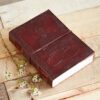 The Sitting Buddha Leather Journal, a closed brown leather piece embossed with its namesake design and secured by a strap, rests gracefully on a wooden surface with small white flowers artfully arranged beside it.