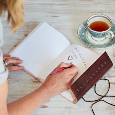 Someone writes "Made with love" using a pink pencil next to an Embossed Leather Journal and a cup of tea on a saucer.