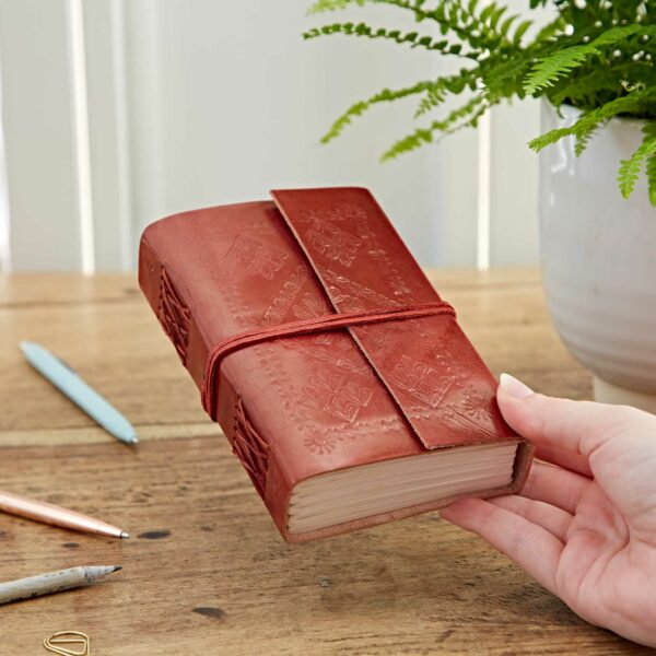A hand holds an Embossed Leather Journal in red, complete with a strap, on a wooden table near a plant and two pens.