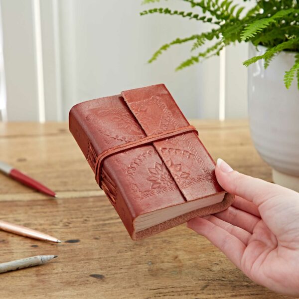 A hand holds the Embossed Leather Journal, a small brown leather-bound notebook with an elastic strap, resting on a wooden table surrounded by pens and a potted plant in the background.