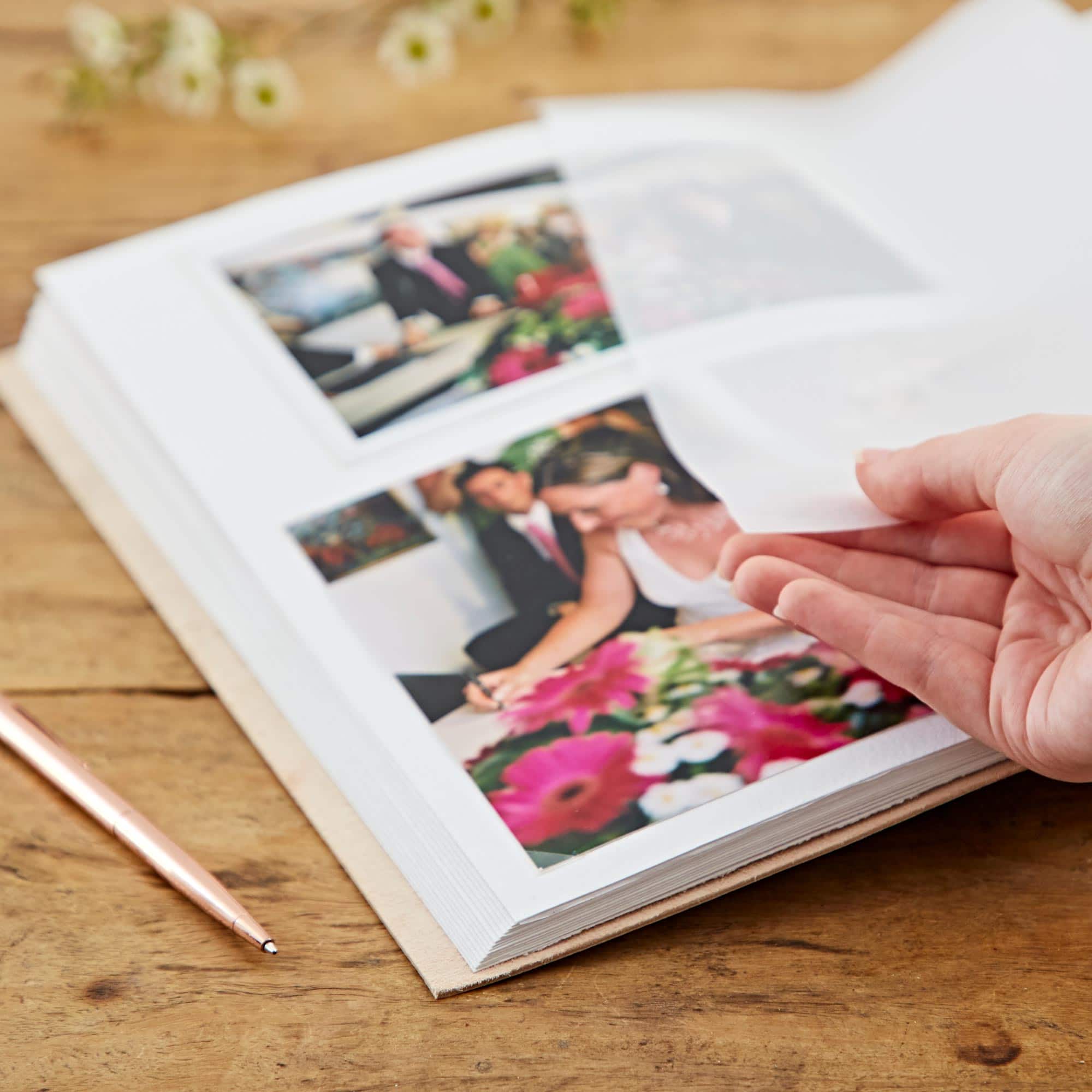 A hand lifts a clear plastic sheet in the Indra Extra Large Embossed & Stitched Leather Photo Album to reveal wedding pictures on a wooden table. A pen lies beside the album.