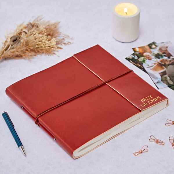 A red leather photo album labelled "Best Grandad" beside a pen, dried flowers, a lit candle, photos, and rose gold paper clips on a white surface.