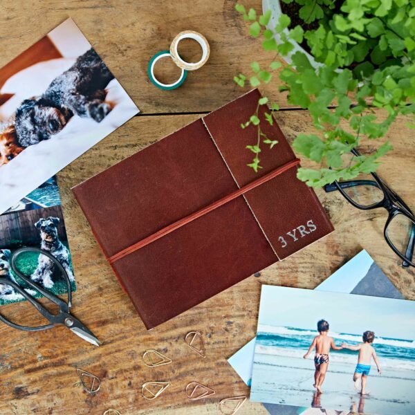 Brown diary labelled "3 YRS" on a wooden table, surrounded by photos, spectacles, scissors, paper clips, a plant, and rolls of tape.