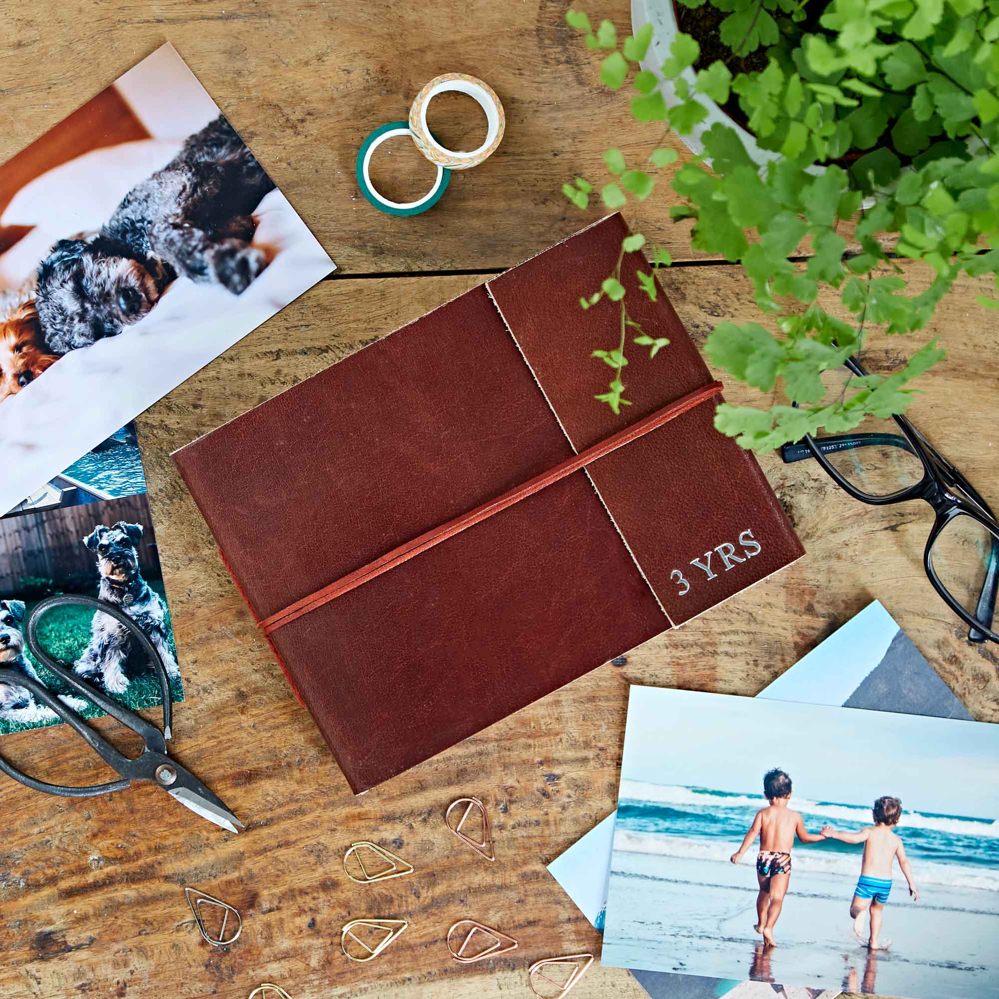 Brown diary labelled "3 YRS" on a wooden table, surrounded by photos, spectacles, scissors, paper clips, a plant, and rolls of tape.
