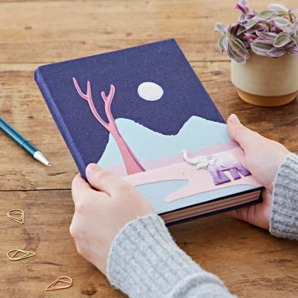 Hands holding a Large Colourful Elephant Dung Notebook with a cover illustration featuring a pink elephant, mountains, a tree, and a moonlit night. A pen, paper clips, and a potted plant are on the wooden table.