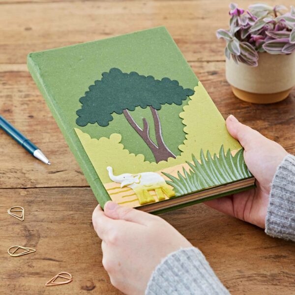 Hands holding a Large Colourful Elephant Dung Notebook with a 3D cutout design featuring an elephant, tree, and grass, on a wooden table with a plant, pen, and paper clips nearby.