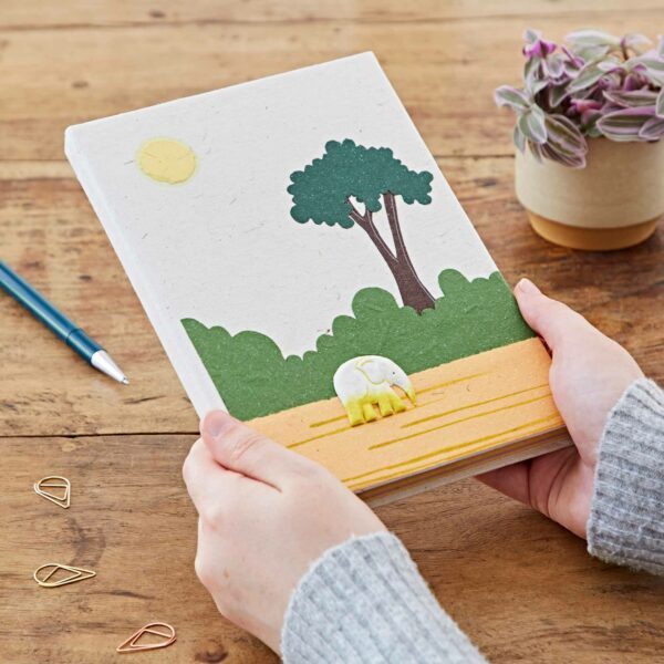 Person holding a Large Colourful Elephant Dung Notebook with an embossed elephant and tree design on the cover, placed on a wooden table with a pencil, paper clips, and a potted plant.