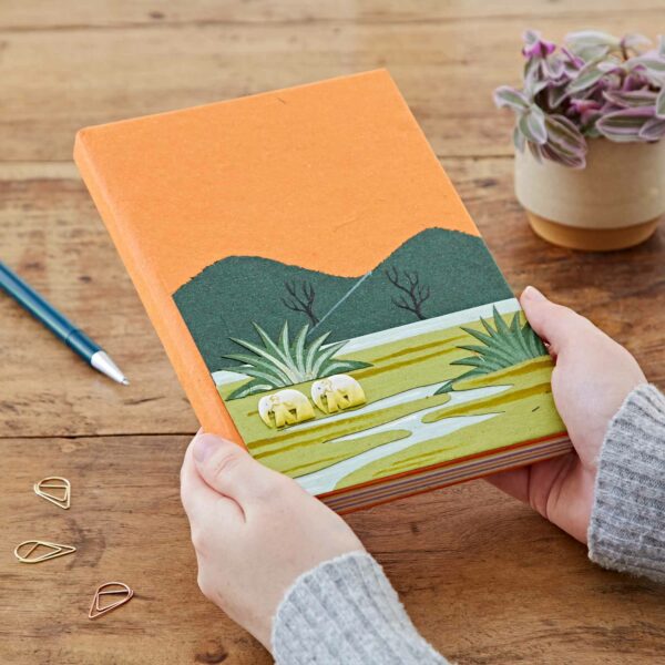 Hands holding a Large Colourful Elephant Dung Notebook with a green and yellow landscape cover design, featuring two small animals. A plant and pen sit on a wooden table in the background.
