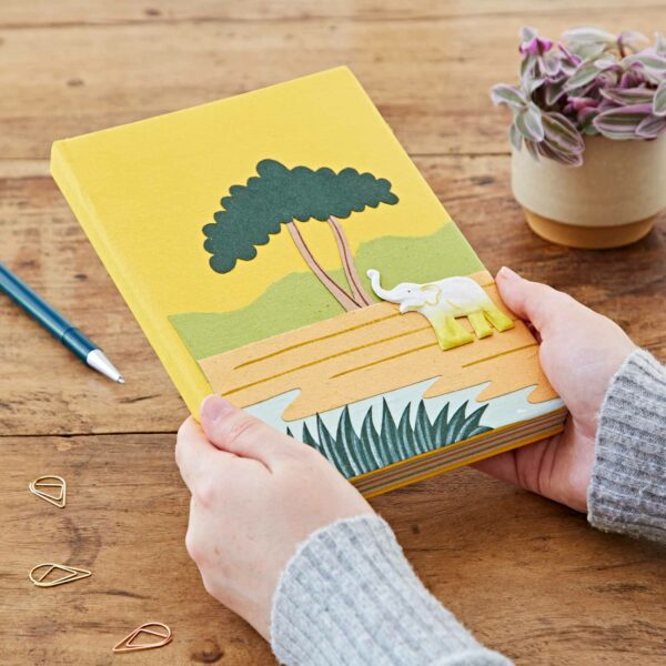 Hands holding a Large Colourful Elephant Dung Notebook with an elephant and tree design on the cover; pencil, paper clips, and a potted plant are on the wooden table.