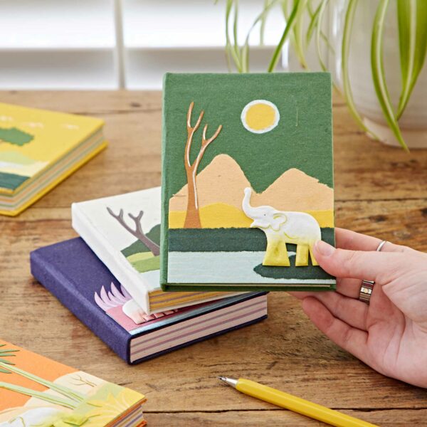 A hand holds a Medium Colourful Elephant Dung Notebook, made from elephant dung, with an elephant and landscape design on the cover. Other colorful notebooks with similar designs are lying on a wooden table. A plant is visible in the background.