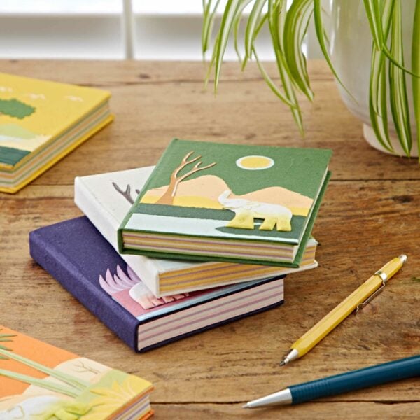 A stack of Medium Colourful Elephant Dung Notebooks with animal illustrations, made from unique elephant dung paper, sits on a wooden table next to a yellow pen, a white pen, and a potted plant.