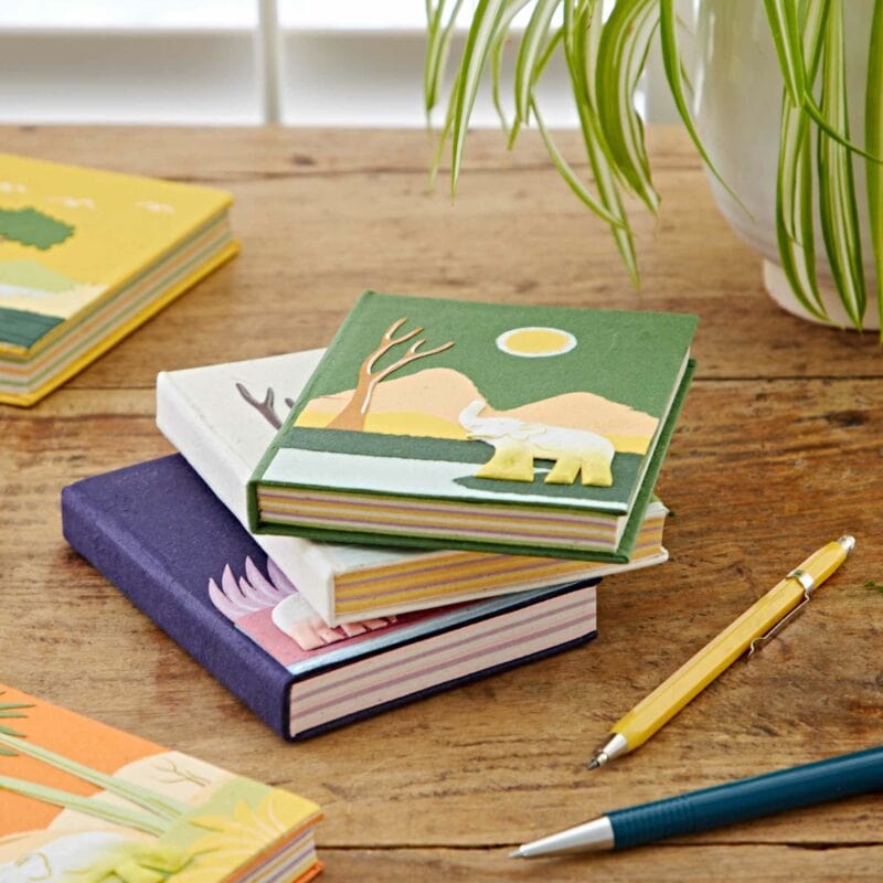 A stack of Medium Colourful Elephant Dung Notebooks with animal illustrations, made from unique elephant dung paper, sits on a wooden table next to a yellow pen, a white pen, and a potted plant.
