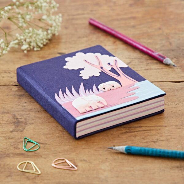 A medium, Colourful Elephant Dung Notebook with a purple cover features a raised design of elephants and trees. Placed on a wooden surface, it is accompanied by a pink pencil, a teal pencil, paper clips, and white flowers nearby.