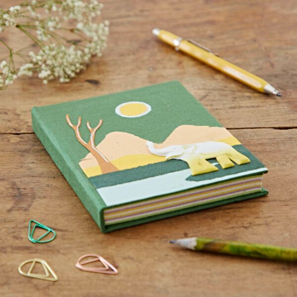 A Medium Colourful Elephant Dung Notebook with an elephant and landscape design on the cover is placed on a wooden surface, surrounded by a yellow pen, green pencil, paper clips, and white flowers.