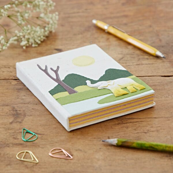 A Medium Colourful Elephant Dung Notebook with a cover illustration of an elephant, trees, and a sun lies on a wooden table. Nearby, there is a yellow pen, a green pencil, some flower stems, and three teardrop-shaped paper clips.