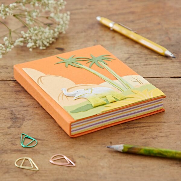 An orange Medium Colourful Elephant Dung Notebook with an elephant and palm tree design on the cover, surrounded by a yellow pen, green pencil, and colorful paper clips on a wooden table.