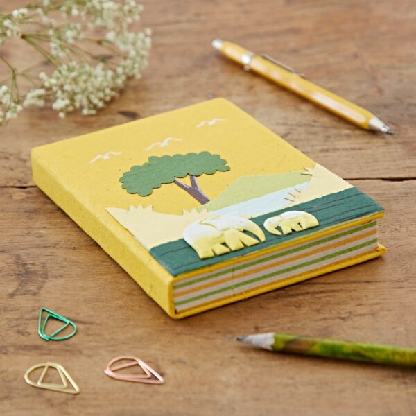 A yellow Medium Colourful Elephant Dung Notebook with a tree and sheep design on the cover is placed on a wooden surface. Nearby are a green pencil, a yellow pen, pink and green paper clips, and white flowers.