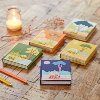 Five colourful notebooks are arranged on a wooden table with a lit candle and two pencils nearby, adding coziness. Among them, one is the "Small Colourful Elephant Dung Notebook," offering an eco-friendly twist with its embossed nature-themed cover.
