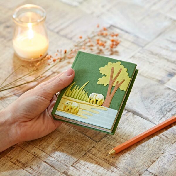 A hand holds a small green notebook with an elephant and tree design. A lit candle, dried flowers, and an orange pencil are on the wooden table.