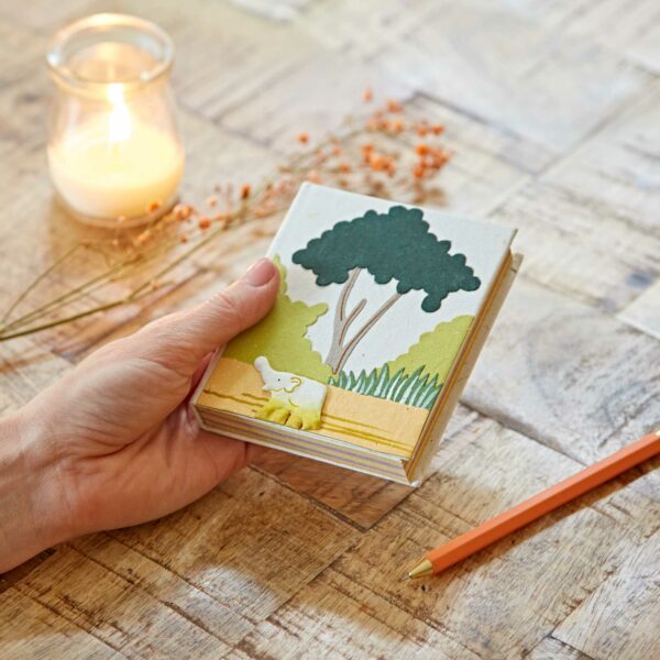Hand holding a small book with an elephant and tree design on the cover. A lit candle and orange pencil are on a wooden table.