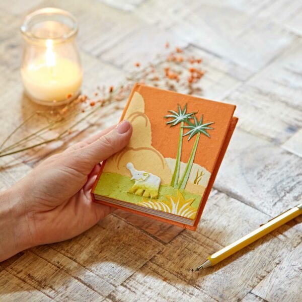 A hand holds a small orange book with an elephant and palm trees design. A lit candle, scattered flowers, and a yellow pen are on the wooden table.