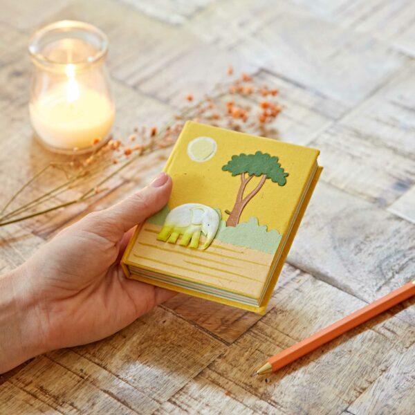 A hand holds a yellow notebook with an elephant and tree design. A lit candle, dried flowers, and a pencil are on the wooden table.