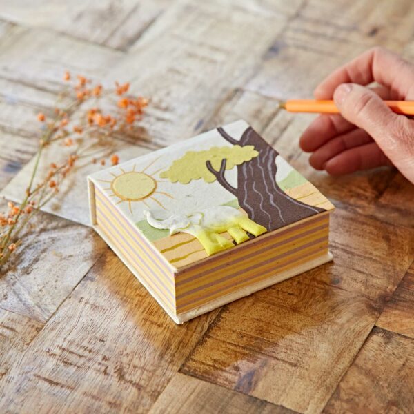 A person writing on a Natural Eco Maximus Elephant Dung Notecube with a sun, tree and elephant design by Paper High.