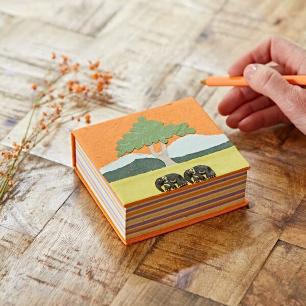 A person writing on a Orange Eco Maximus Elephant Dung Notecube with a tree and elephant design by Paper High.