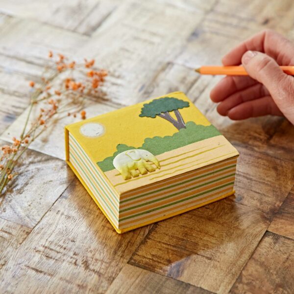 A person writing on a Yellow Eco Maximus Elephant Dung Notecube with a sun, tree and elephant design by Paper High.