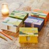 Five vibrant, nature-themed boxes featuring animal designs, including a Colourful Elephant Dung Memo Box, are displayed on a wooden table beside a candle and two pencils.