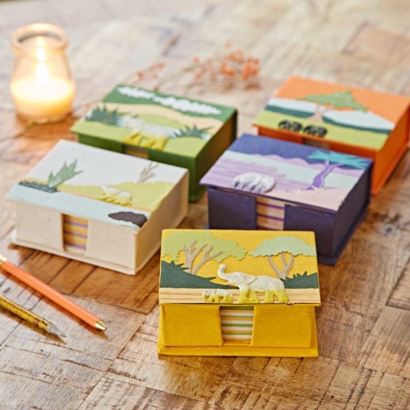 Five vibrant, nature-themed boxes featuring animal designs, including a Colourful Elephant Dung Memo Box, are displayed on a wooden table beside a candle and two pencils.