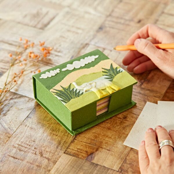 A hand holding an orange pen is next to a green box with a decorative elephant design on top, placed on a wooden table with dried flowers.