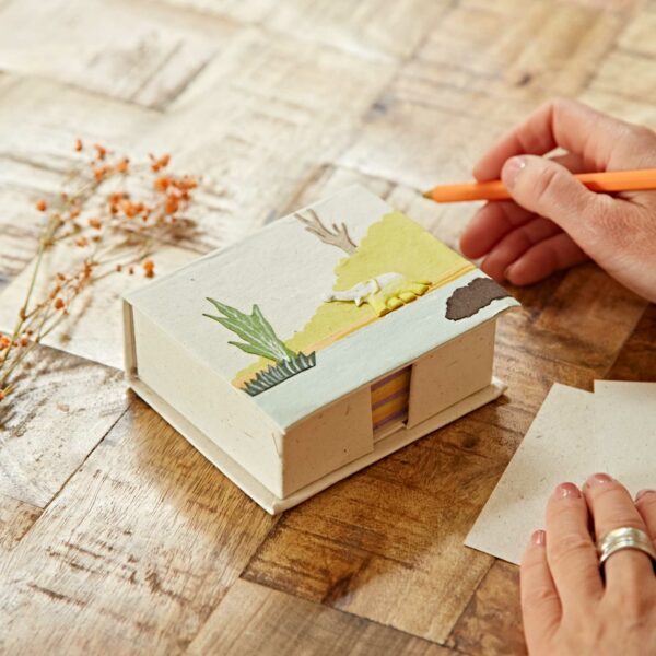 A person decorates a small box with a nature-themed illustration on a wooden table. Orange pencil and dried flowers are nearby.