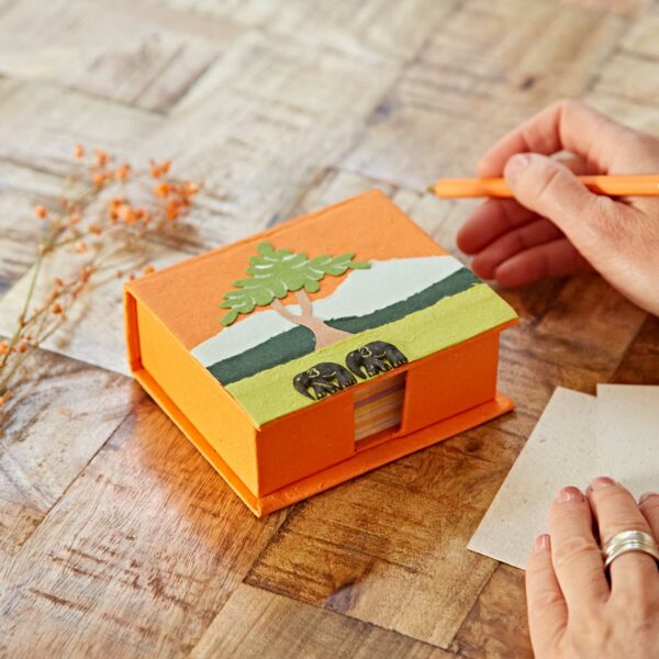 Person writing on paper next to an orange box shaped like a house with a tree design, on a wooden table with decorative orange twigs.