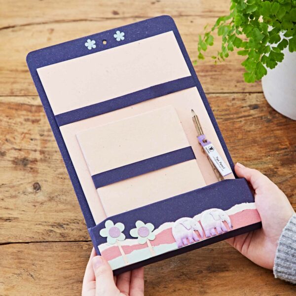 Hands holding a decorative, colourful clipboard with pastel stationery, a pen, and a design featuring flowers and elephants. A potted plant is partially visible in the background, reminiscent of a Colourful Elephant Dung Letter Writing Set.