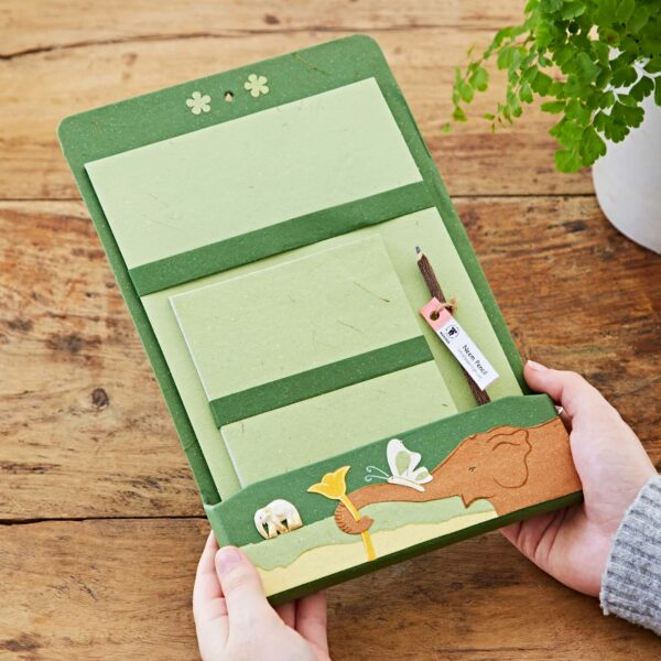 A person holding a green Colourful Elephant Dung Letter Writing Set with elephant-themed designs, papers, a notepad, and a pen on a wooden table next to a plant. The colourful elephant dung stationery adds a unique touch.