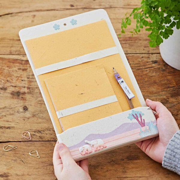 Hands holding a beige Colourful Elephant Dung Letter Writing Set on a wooden table. The set includes envelopes, a pencil, and has a nature-themed decorative band with illustrations of dogs and a tree. It’s the perfect blend of practicality and charm, ideal for those who appreciate unique touches like Colourful Elephant Dung paper.