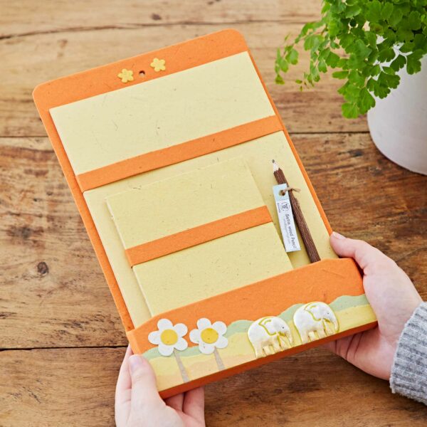 Two hands holding a vibrant Colourful Elephant Dung Letter Writing Set with an orange and cream-colored stationery, featuring a flower and Colourful Elephant Dung design, placed on a wooden surface next to a green potted plant.