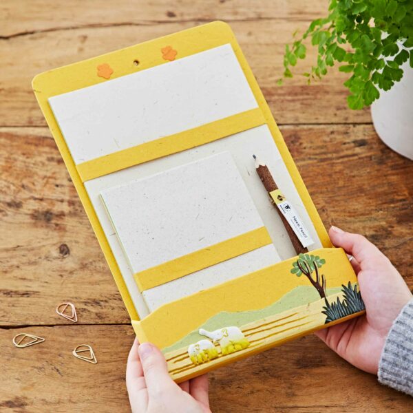 Hands holding a Colourful Elephant Dung Letter Writing Set featuring yellow borders, a pencil, and an illustration of a tree and sheep on a farm, placed on a wooden table with paperclips and a potted plant.