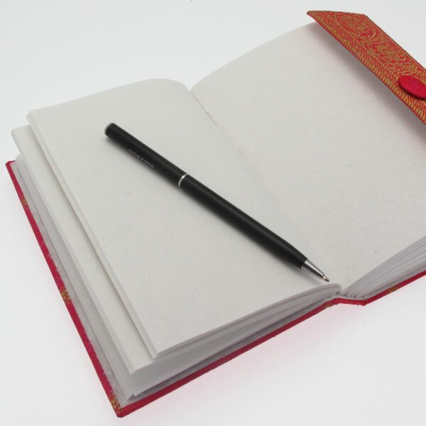 A black pen rests on an open Medium Sari Fabric Journal, featuring blank pages. The notebook is adorned with a red cover and fastener, inviting thoughts to flow freely.