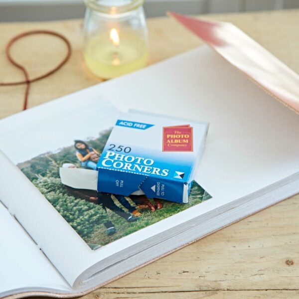 An open photo album on a wooden table shows a photo and a box of 250 Clear Self-Adhesive Photo Corners resting on its pages, while a glowing candle in the background creates a warm, inviting atmosphere.