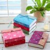 Large Sari Fabric Journals with decorative closures are stacked on a wooden table beside a green plant, candle, and pen.