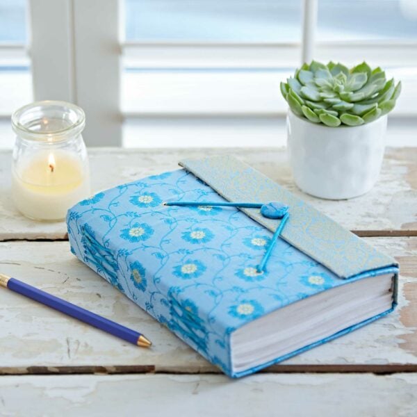 A blue floral-patterned journal with a button closure is on a wooden table. A blue pencil, a lit candle, and a small potted succulent are nearby.