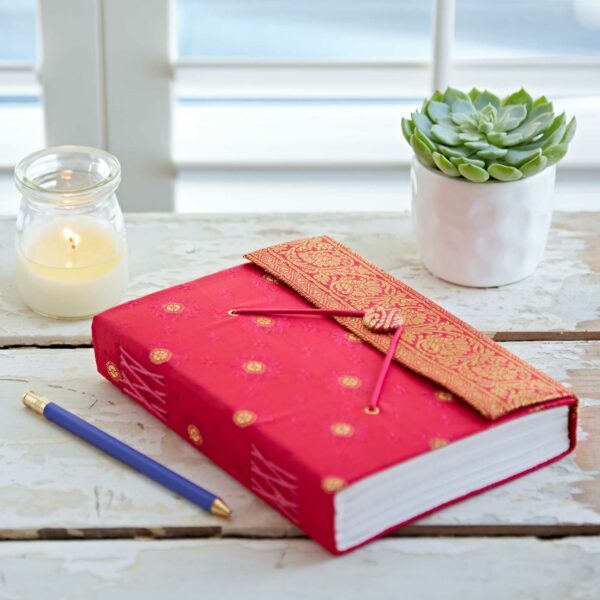 A red fabric-bound journal and a blue pen on a wooden table with a lit candle and a small potted succulent in the background.