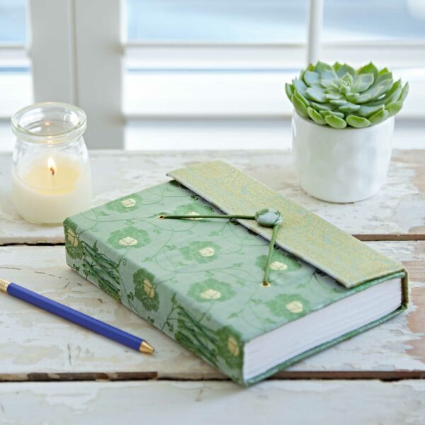 A green floral-patterned journal and a blue pencil lie on a table near a candle in a glass jar and a small pot with a succulent.